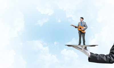 Businessman on metal tray playing acoustic guitar against blue sky background