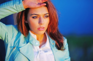Young charming girl in turquoise jacket, lit by bright rays of setting sun on a blurred background of a beautiful evening sky and the greenery of Park, closeup