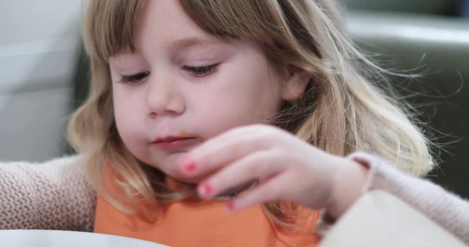 4k video close up shot. Three years old blonde baby with orange bib eating pizza piece with her hands, nails red painted, sitting in green chair in table of restaurant
