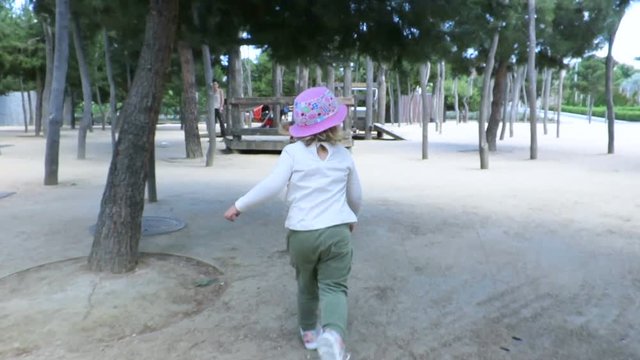 two years old blonde child with green trousers, white shirt and pink hat playing and running in urban playground park in Madrid city, Spain
