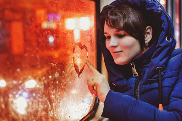 Young single woman in blue jacket sitting in an empty tram and paints on glass heart.