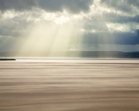 Crepuscular rays through a stormy sky while shifting sands create a cloud underfoot as wind whistles across the beach, West Kirkby, Wirral