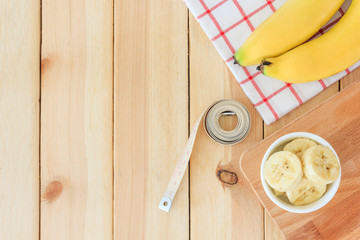 Two bananas and banana slices in white bowl with measuring tape on wooden table background, Top view with copy space and text.