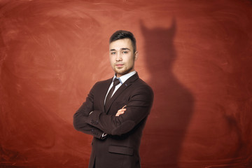 Businessman in suit standing with his arms folded, he is casting shadow of the devil on the rusty orange wall behind him.