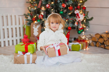 Fototapeta na wymiar Little beautiful girl sitting near the Christmas tree with gifts in his hands. in a white sweater and warm headphones