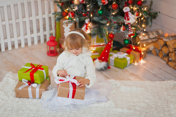 Fototapeta na wymiar Little beautiful girl sitting near the Christmas tree with gifts in his hands. in a white sweater and warm headphones