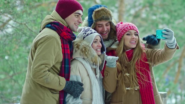 Two young men and two beautiful women standing together in winter forests and recording video with smart phone: they talking at camera, smiling and waving