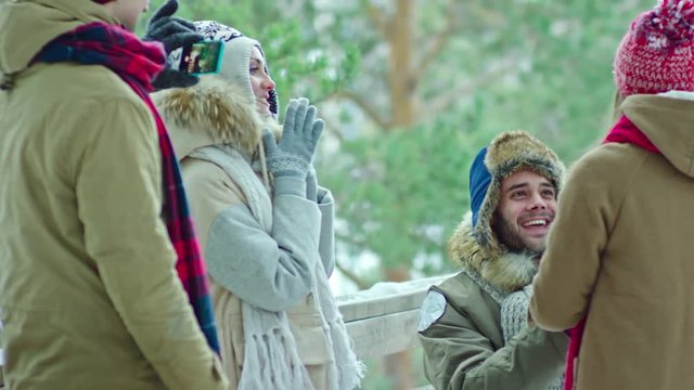 Young man standing on one knee and asking girlfriend marry him while their friends making video with smart phone. She answering yes, they happily smiling and embracing at winter day outdoor