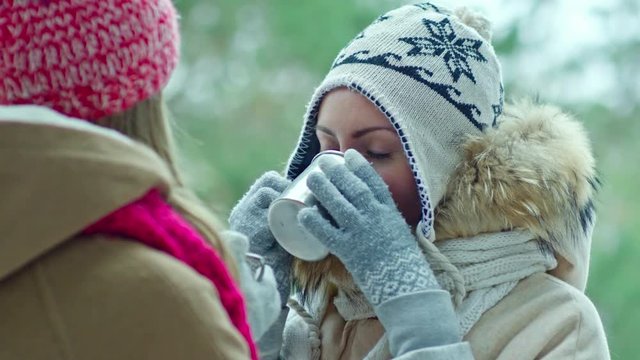 Closeup of beautiful woman with blue eyes drinking hot tea from thermo cups and speaking with her friend at winter day in the forest