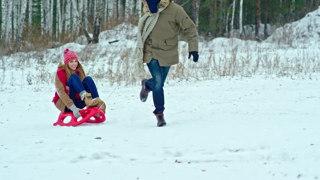 Man running in park at winter day and pulling his laughing girlfriend on sledge in slow motion. He slipping on snow and woman falling down