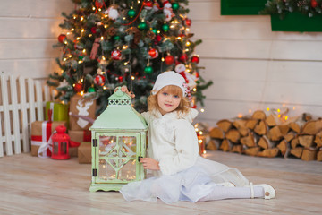 Obraz na płótnie Canvas Little beautiful girl in a white hat and clothes sits near Christmas tree