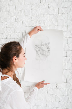 Artist hanging her pencil portrait on wall. Close-up of attractive woman decorating white brick side in her workshop with sketch. Art, creativity, hobby, talent,drawing, relax concept