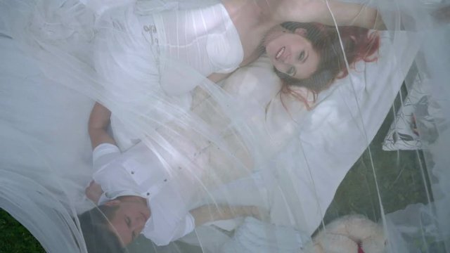Top view of love couple relaxing on white fabric behind transparent veil in park. Romantic holiday. Closeup of two people lying in romantic decoration. Romantic couple resting outdoor