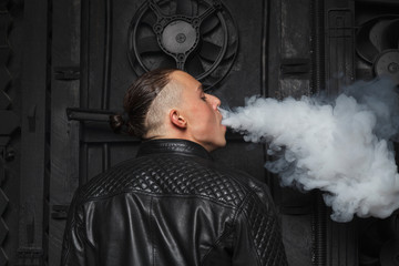 Fototapeta na wymiar Fashionable and stylish man standing on black background with details and smoke E-cigarette in a leather black jacket, gray smoke releasing