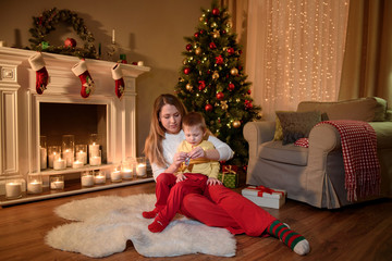 A young mom with her son on her knees sitting on a furry carpet and opening a chocolate sweet for him.