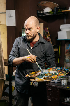The artist looks thoughtfully at his painting with a brush in his hand. Art process
