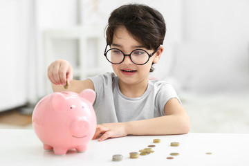 Obraz na płótnie Canvas Portrait of cute little boy in glasses putting coin into a piggy bank on blurred background