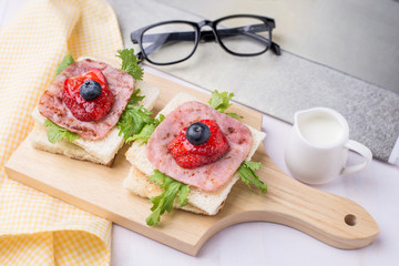 Healthy breakfast with toasted bread, milk, ham, vegetables, strawberry, jam and blueberry topping on wooden plate. Top view with white wooden table background.