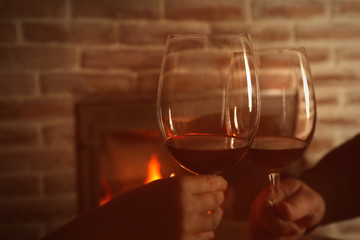 Hands of couple clinking glasses with red wine against blurred fireplace