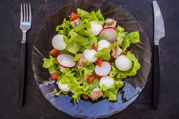 Salad of lettuce, radishes, crackers, tomato and cheese on a transparent dark Circassian plate. Wooden table