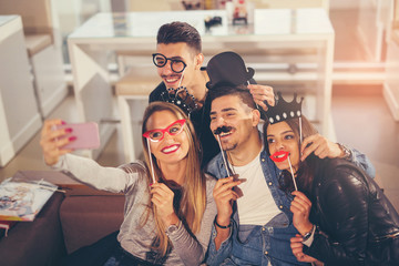 Young friends having a great time in restaurant holding artificial mustache and lips stick making selfie photo