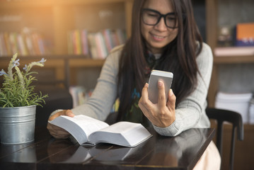 Fototapeta premium Beautiful woman wearing glasses and using smartphone and reading a book in the library.She smiled, she was happy selfie by phone.
