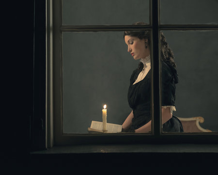 Retro victorian girl reading book by candlelight behind window.