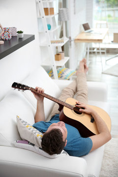 Young man playing guitar and lying on the sofa in light room