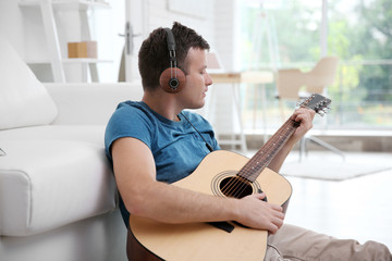 Young man with headphones playing guitar and sitting on the floor in the room