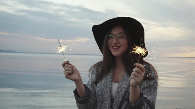 Pretty happy woman rejoicing with sparklers on the sea coast at sunset slow motion