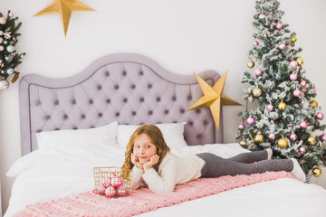Obraz na płótnie Canvas Portrait of cute happy little girl on Christmas morning in home interior. Child paying with holiday decoration while laying on bed. Horizontal color photo.