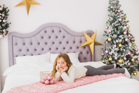Portrait of cute happy little girl on Christmas morning in home interior. Child paying with holiday decoration while laying on bed. Horizontal color photo.