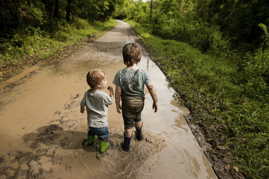 Rear view of messy brothers walking in dirty puddle on road