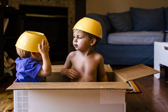 Playful brothers with bowls on head sitting in cardboard box at home