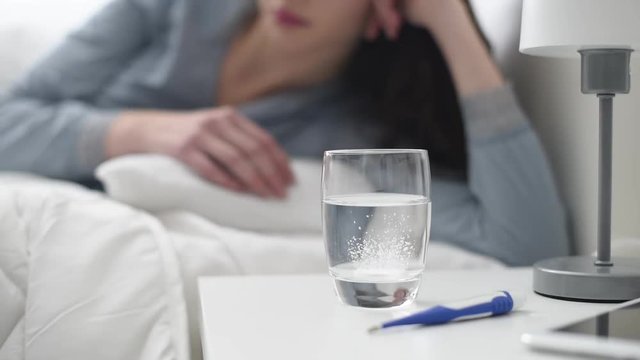 Sick woman lying in bed, she is putting an effervescent fizzing tablet into a glass of water, medicine and healthcare concept