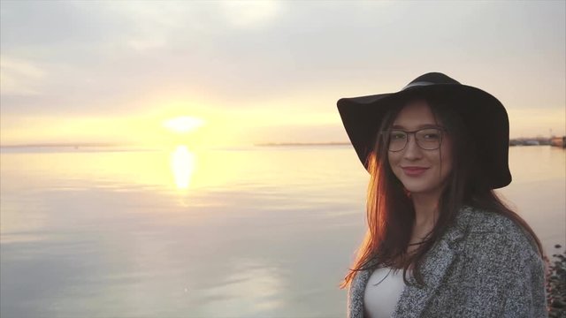 Young pretty smiling woman in black hat and glasses near the sea at sunset HD