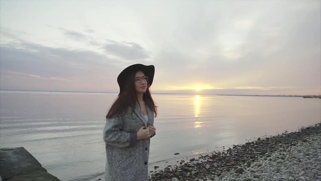 Young pretty laughing and smiling woman in black hat and glasses near the sea at sunset