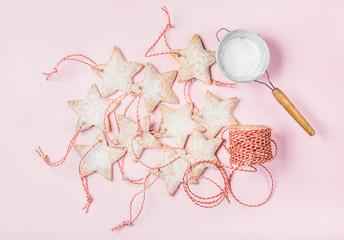 Christmas homemade gingerbread star shaped cookies with sugar powder in sieve and red decoration rope over light pink background, top view, horizontal composition