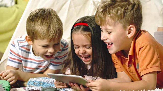 Lockdown of laughing little children lying on floor and watching something funny on tablet