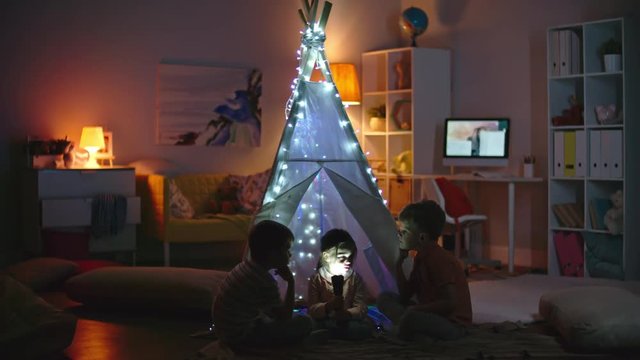 Adorable girl holding flashlight in her face and telling scary stories to little boys sitting in dark room near teepee decorated with fairy lights
