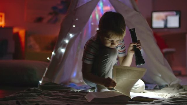 Little boy with flashlight sitting in his room near teepee decorated with fairy lights and looking at book