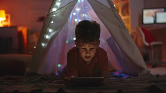 Little boy lying in dark in his teepee decorated with fairy lights and watching something on tablet