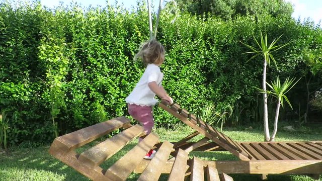 two years age blonde cute baby with white shirt jumping and playing on wooden hammock in green garden plants
