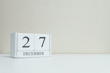 Closeup white wooden calendar with black 27 december word on blurred white wood desk and cream color wallpaper in room textured background with copy space , selective focus at the calendar