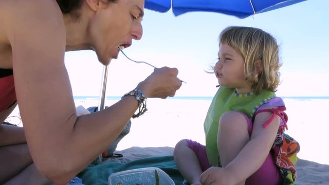 little baby and mother sitting down of parasol at beach sharing eating salad from tupperware