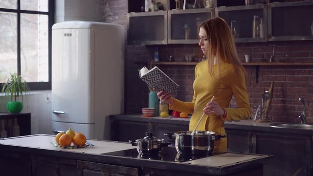 Elegant adult woman cooking and reading book in the contemporary kitchen. Young lady boiling soup at home. Caucasian woman with long hair wearing in stylish dress.