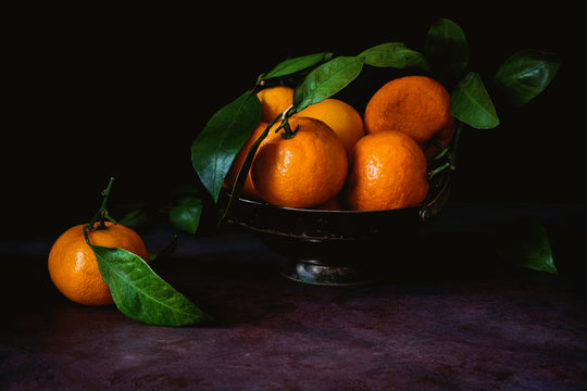 Still life of fresh tangerines with green leaves in vintage metal bowl. Toned image, low key technique (chiaroscuro). Close up view.