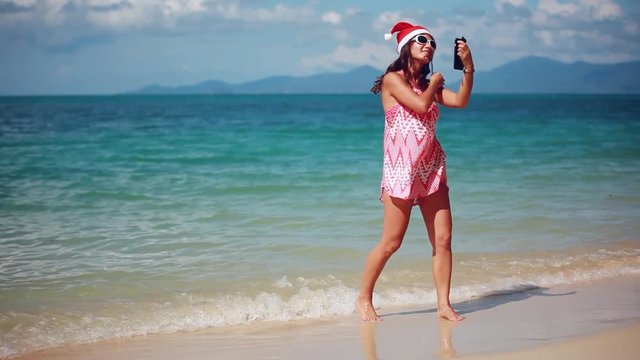Christmas young slim woman in red santa hat and sunglasses taking picture self portrait on smartphone at beach over sea background on Koh Samui. Thailand. 1920x1080