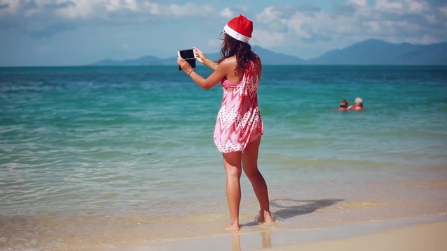 Christmas young smiling woman in red santa hat and sunglasses taking picture self portrait on smartphone at beach over sea background on Koh Samui. Thailand. 1920x1080