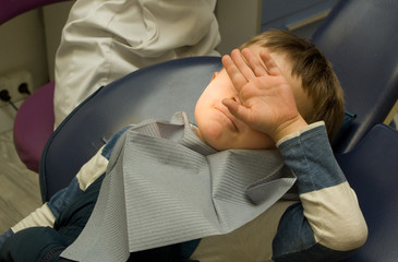 Five y.o. boy doesn't want to make a stock photo sitting in the dentist chair under the medical lamp light and closing face by his arm - 130609365